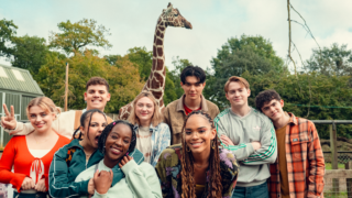 Heartstoppers group shot of all the kids at the zoo with Charlie leaning on Nick's shoulder and a giraffe standing tall in the background